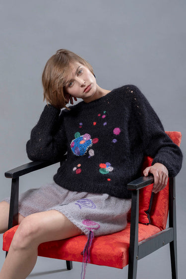 Woman sitting on red chair wearing black mohair sweater and grey mohair shorts. The sweater has colourful abstract star-like embroideries adorning it.